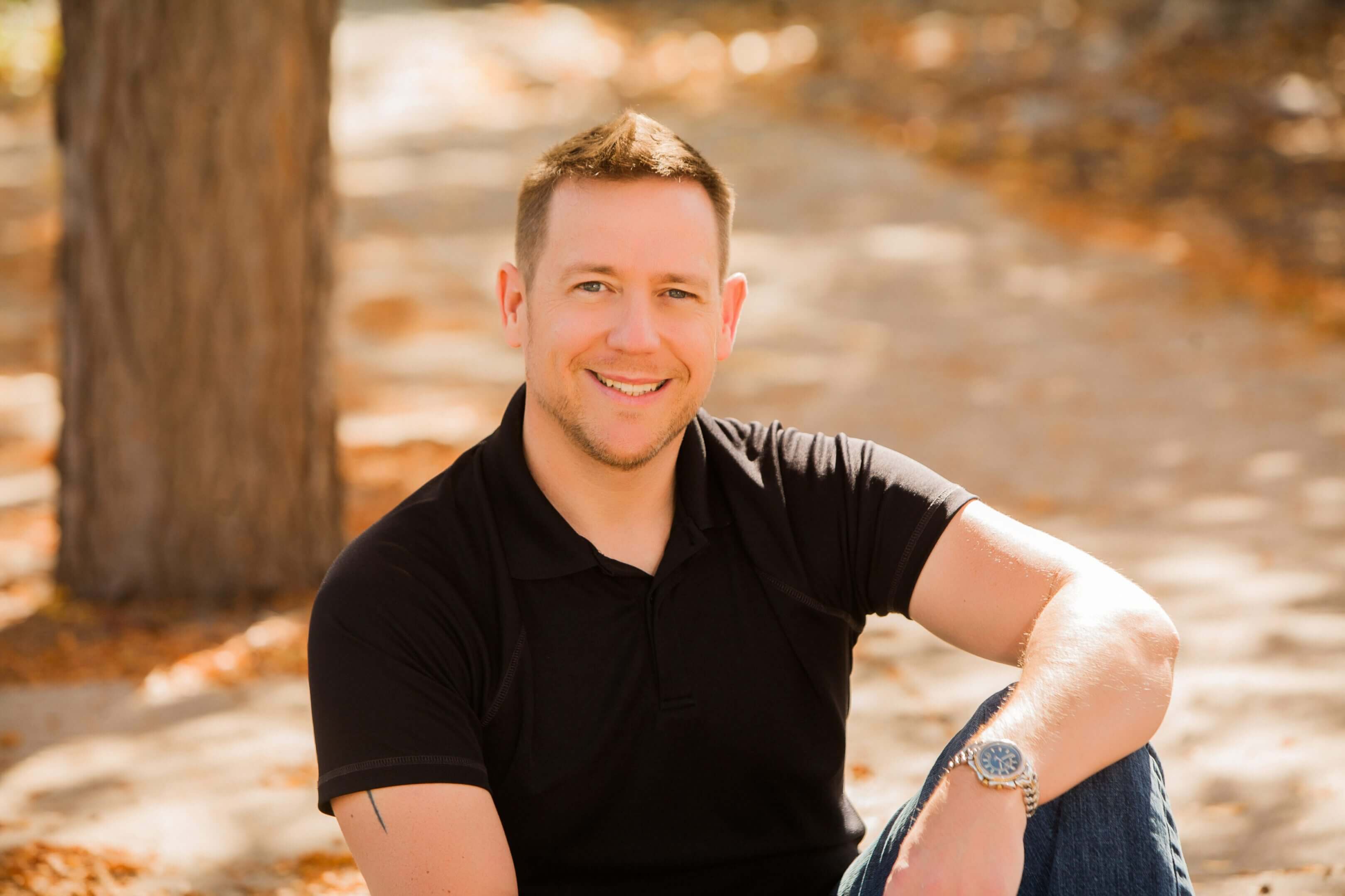A man in a black shirt sitting outdoors with a smile.