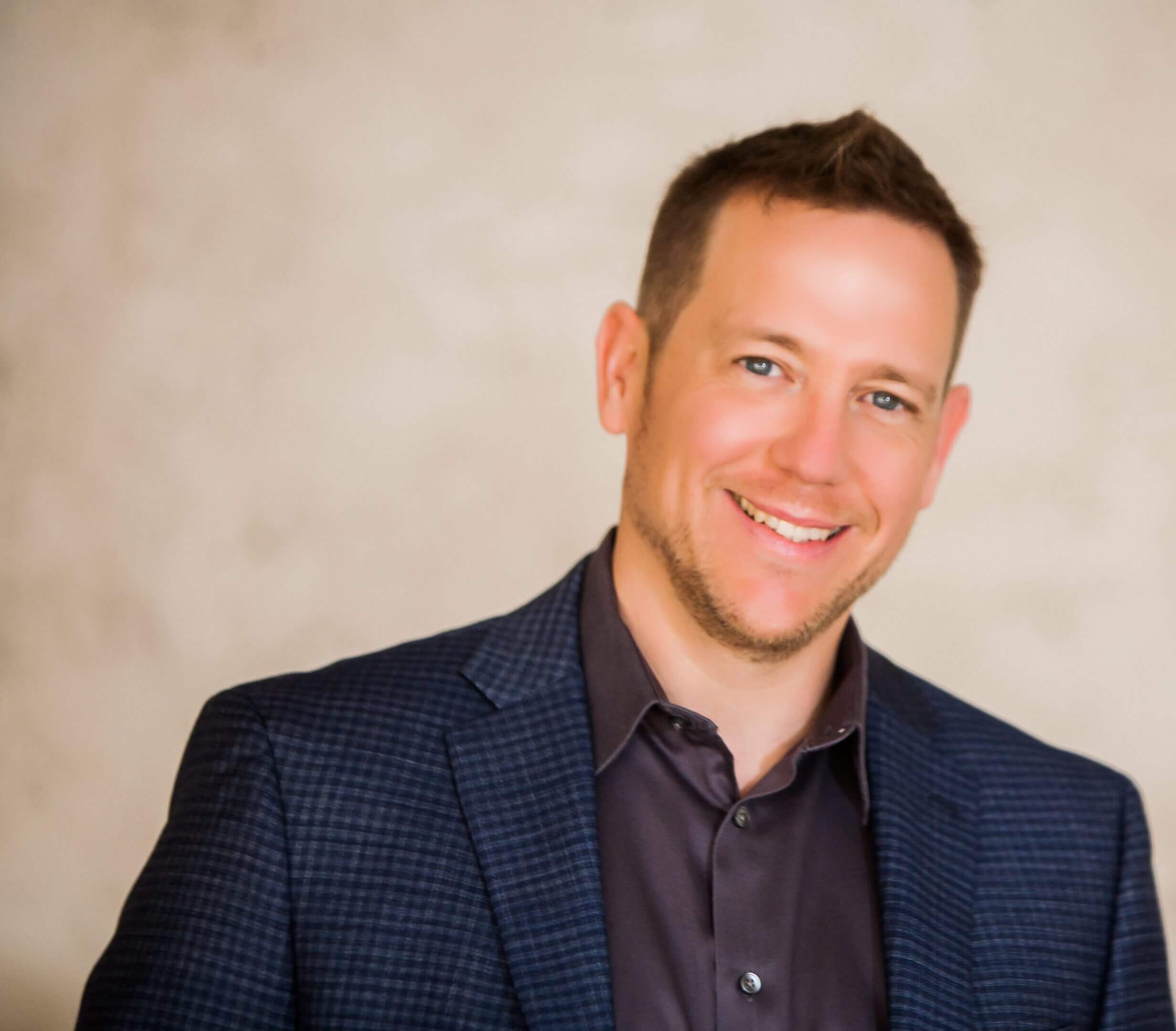 Smiling man in a dark blazer and shirt against a neutral background.